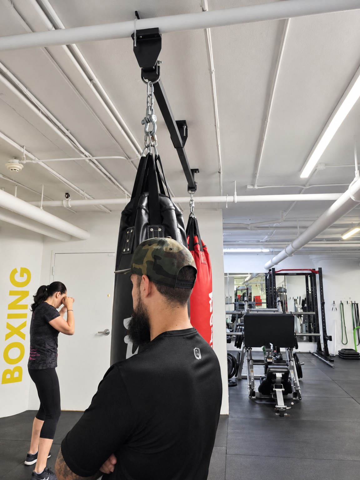 Gianluca observing a woman practising boxing in a real gym, beside the heavy bags.