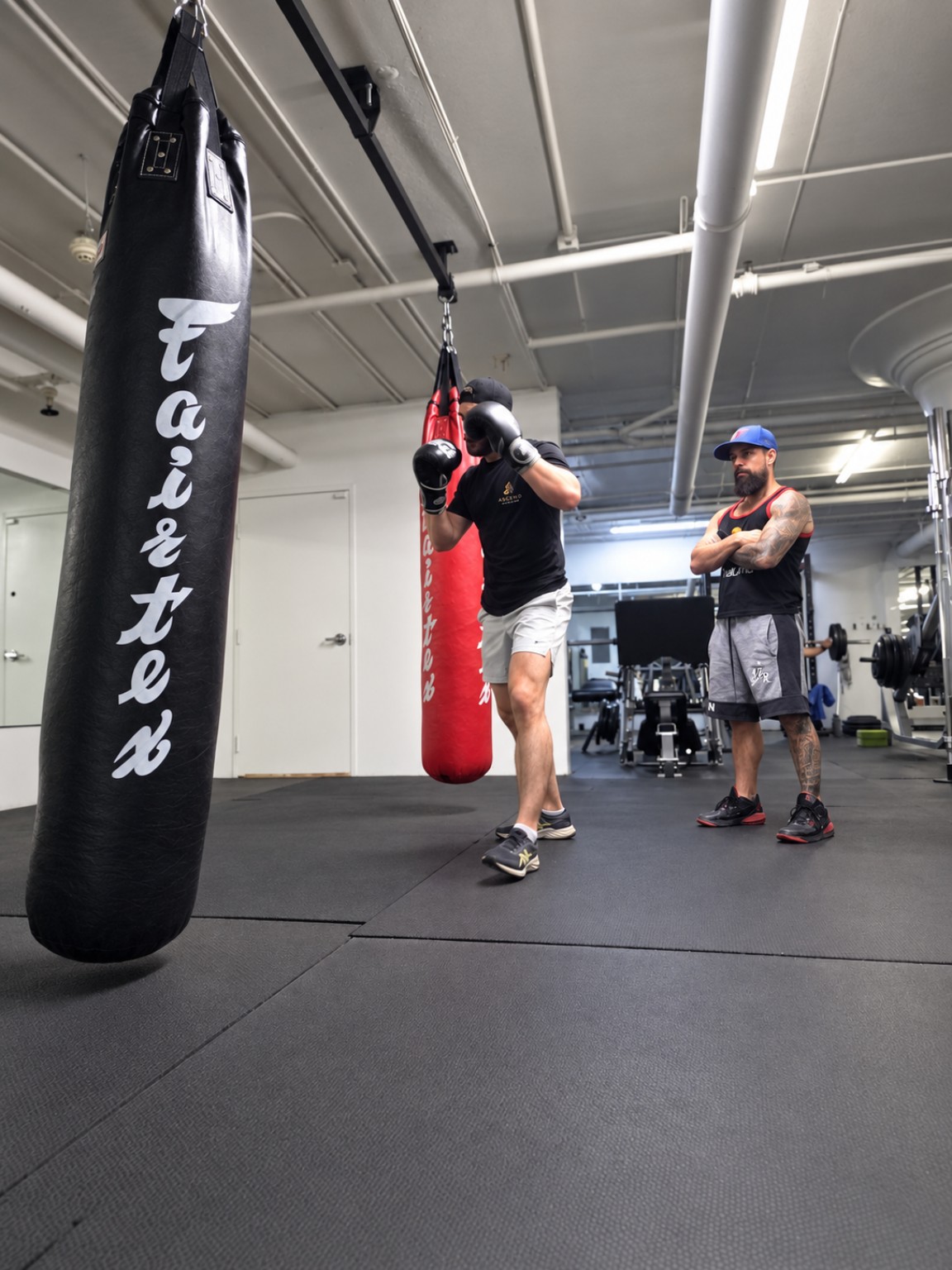 Gianluca coaching a client during striking practice, beside black and red Fairtex heavy bags.